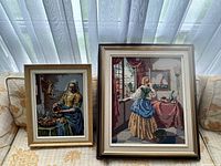 Two framed needlepoint artworks displayed on a sofa, showing a woman pouring liquid and another standing by a table