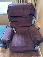 Full view of a purple fabric lift recliner chair with padded armrests and a cushioned backrest. Remote control is visible on the right armrest.