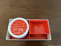 Top view of novelty coaster with Irish toast quote and small figure illustration, inside the white box with red compartments.