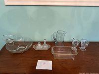 Wide shot showing all glass items on a wooden surface against green wall including rectangular baking dish, pitcher, small vases, double inkwell, and glass dish with bowl and spoon.