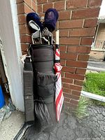 View of entire golf bag in black cloth with shoulder strap and several pockets, with multiple golf clubs and an umbrella attached.