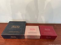 Three Trivial Pursuit game boxes - Master Game Genus Edition (large, black), Silver Edition (beige), and Easy Wedges Edition (red) arranged on a wooden surface