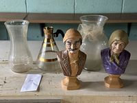 Five vintage decor items including three glass vases and two ceramic bust figurines on a wooden surface.