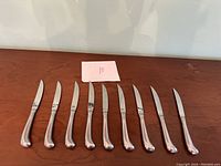 Nine kitchen knives with curved polished metal handles arranged on wooden surface.
