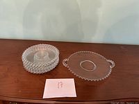 Set of clear vintage glass dishes including stacked round plates and an oval serving tray with beaded edges, placed on wooden surface against white wall.