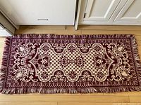 Full view of rectangular burgundy and cream wool jacquard mat with fringe on wooden floor.