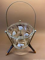 Front view of glass ice bucket in metal tripod frame with detailed gold and white leaf painted design on the glass bowl.