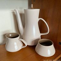 Full view of coffee pot, cream pitcher, and sugar bowl on wooden surface against wall background.