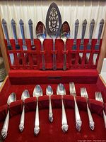 Front view of the cutlery set laid in a wooden display box with red velvet lining. Visible are the 8 knives stored upright in slots, and forks and spoons organized below. Two large serving spoons and one pierced fish server spoon are mounted in the box lid.