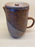 Front view of lidded pottery jug showing speckled rust, sandy, and chocolate glaze.