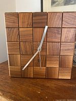 Front facing photo showing teak checkerboard clock face with 25 wooden cubes and matte silver arms in natural indoor lighting.