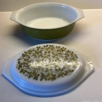 Open vintage Pyrex casserole dish showing olive green handle and white interior with matching lid featuring green foliage design laid in front.