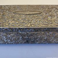 Close-up photo showing detailed raised repousse floral and foliage pattern on the metal surface of the jewellery box.
