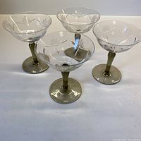 Four vintage crystal champagne bowls with clear etched floral pattern bowls and smoky grey stems and feet on white surface.
