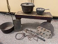 Wide view of antique cast iron cookware and hooks arranged on wooden bench and ground, showing all items in the lot.