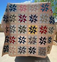 Full view of antique quilt showing 25 patchwork star blocks in a 5x5 grid, with varied colors and patterns, including blues, tans, reds and yellows, bordered by a floral print fabric.
