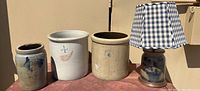 Front view of three antique stoneware crocks of different sizes and a stoneware lamp with a blue gingham lampshade arranged on a surface.