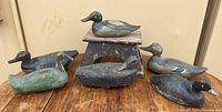 Photo showing six vintage wooden duck decoys arranged on a wooden surface with a small wooden pedestal in the background. Wear and paint chipping visible on the decoys.