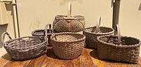 Photo of five old woven baskets on a wooden surface against a beige wall. The baskets vary in shape and size, some with handles and some without.