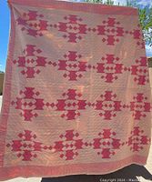 Full view of the quilt showing the repeating geometric pattern in red and pink on a light background, hung outdoors.