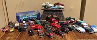 Wide view of approximately 30 model cars arranged on a wooden surface including boxed and loose items in varied colors.