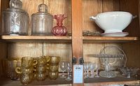 Photo of vintage glassware and crystal arranged on wooden shelving, showing various glass jars, pressed glass cups, crystal stemware, and ceramic bowl set.