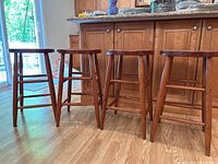 Four cherry finish wooden bar stools shown side by side from front and side angles on hardwood floor near kitchen cabinets.