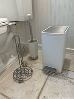 Photo of bathroom items: spiral toilet paper holder, toilet bowl brush with holder, and Kohler pedal garbage pail on tiled floor.