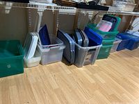 Wide angle photo showing various plastic storage containers lined up on a floor beneath a shelf.