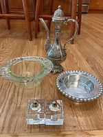 Photo showing a decorative metal teapot with embossed patterns, a clear glass bowl with scalloped edge, a metal bowl with beaded rim, and a pair of square glass salt and pepper shakers with metal tops, all placed on a wooden floor.