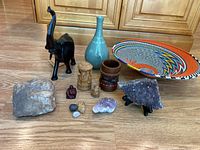 Photo showing the full assortment of travel souvenirs including wooden elephant figurine, ceramic vase, woven basket bowl, carved figures, bracelets, and mineral specimens on a wooden floor.
