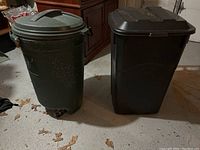 Photo of two garbage pails, one round dark green with lid and wheels, and one black rectangular with flat lid, placed on a concrete garage floor near cabinetry.