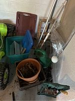 Overview of various garden tools and pots including frog watering can, plant pot with soil, gardening hand tools, rake, plastic bins, and metal poles