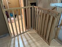 Photo showing a wooden gate with two foldable panels connected by metal hinges. The gate is made of vertical wooden slats and horizontal rails and is displayed standing on a wooden floor.