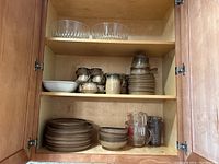 Open wooden kitchen cabinet with three shelves containing brown ceramic dishes and bowls, two large clear glass bowls, and measuring cups in glass and plastic.