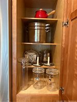 Kitchen cabinet containing stainless steel pot with two steamer inserts, red fondue pot, Cuisinart food processor parts, and three plastic storage jars