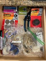 Overhead photo of a wooden drawer containing assorted household repair and utility items: boxed furniture repair markers, multiple rolls of Scotch tape, rubber bands, cutters, pens, carabiners.
