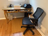 Photo of wood desk with glass top, two drawers open showing office supplies, black HP OfficeJet 4650 printer on top, and black ergonomic mesh office chair with armrests beside desk.