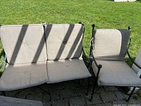Photo of the outdoor wrought iron loveseat and armchair with beige cushions on a patio with green grass background.
