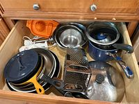 Drawer with multiple pots and pans, orange citrus juicer, stainless steel bowls, a box grater, blue saucepan with glass lid, yellow cast iron skillet, and a white immersion blender
