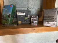 Four protective covers displayed on wooden shelf: green tarp in packaging on left, black patio chair cover box in middle-left, Crivit smartphone bike pouch in middle-right, and grey bicycle cover in packaging on right.