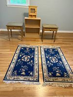 Overview of both end tables, bookshelf, mini hutch, and two blue floral area rugs laid out on wooden floor beneath a window.