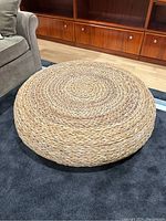 Photo of large, low, round braided wicker coffee table placed on dark carpet near sofa and wooden cabinetry