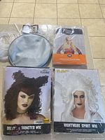 Four Halloween wigs in original sealed packaging displayed on a floor, including medieval witch, deluxe character with cat ears, nightmare spirit white, and a silver/grey wig with round bag.