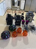 Full view of all items on kitchen counter: 3 black metal lanterns, 2 black pumpkin candle holders, 3 orange glass pumpkin candle holders, metal Happy Halloween sign, 2 purple candle holders, metal owl and bat tealight holders