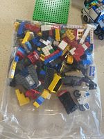 Clear plastic bag with assorted LEGO bricks in various colors including red, blue, yellow, black, and white, tightly packed and laid on a flat surface with a green LEGO baseplate in background.