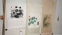 Three Asian paper scrolls hanging side-by-side on a wall, showing panda, bamboo, and landscape artwork