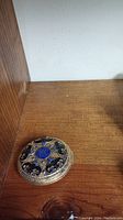Closed round Asian pill box with gold-tone intricate filigree and blue and black inlay on wooden surface.