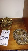 Two brass lion head door knockers placed side by side on a shelf, showing details and size difference.