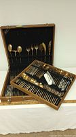 Full view of the open wooden case containing the bronze flatware and serving pieces organized inside, showing spoons, forks, and knives in rows with black felt lining.